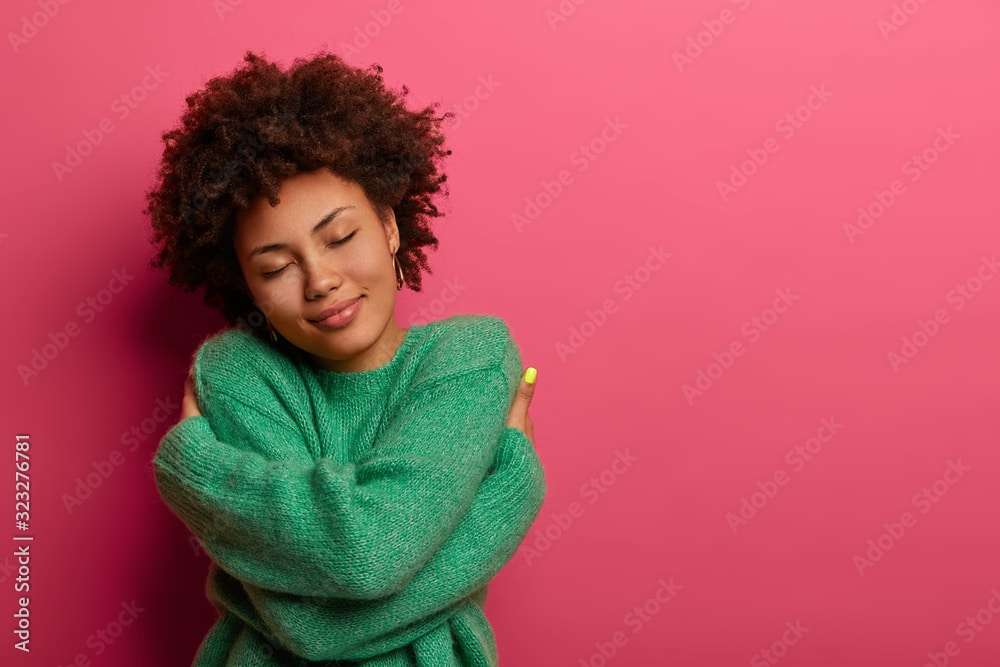 Cheerful young romantic woman expresses self love and care, tilts head and smiles gently, wears green oversized jumper, embraces own body, closes eyes, stands in studio against pink background