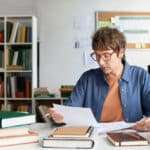 Person analyzing documents and notes in a well-organized library filled with books and plants. Engaging in a detailed study session while seated at a desk