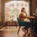 Young Handsome Man Working from Home on Desktop Computer in Sunny Stylish Loft Apartment. Creative Designer Wearing Cozy Yellow Sweater and Headphones. Urban City View from Big Window.
