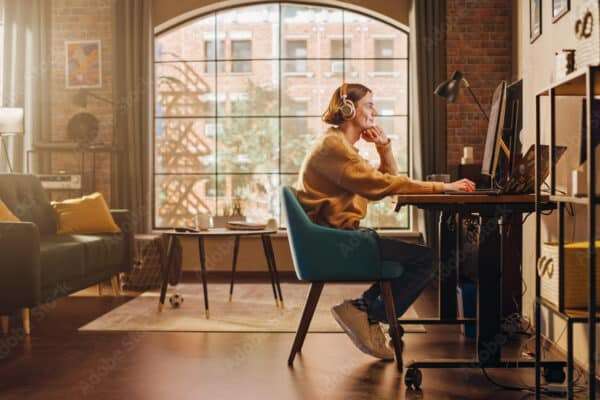 Young Handsome Man Working from Home on Desktop Computer in Sunny Stylish Loft Apartment. Creative Designer Wearing Cozy Yellow Sweater and Headphones. Urban City View from Big Window.