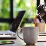 A hand pouring steaming coffee in to a cup on a work desk when work from home